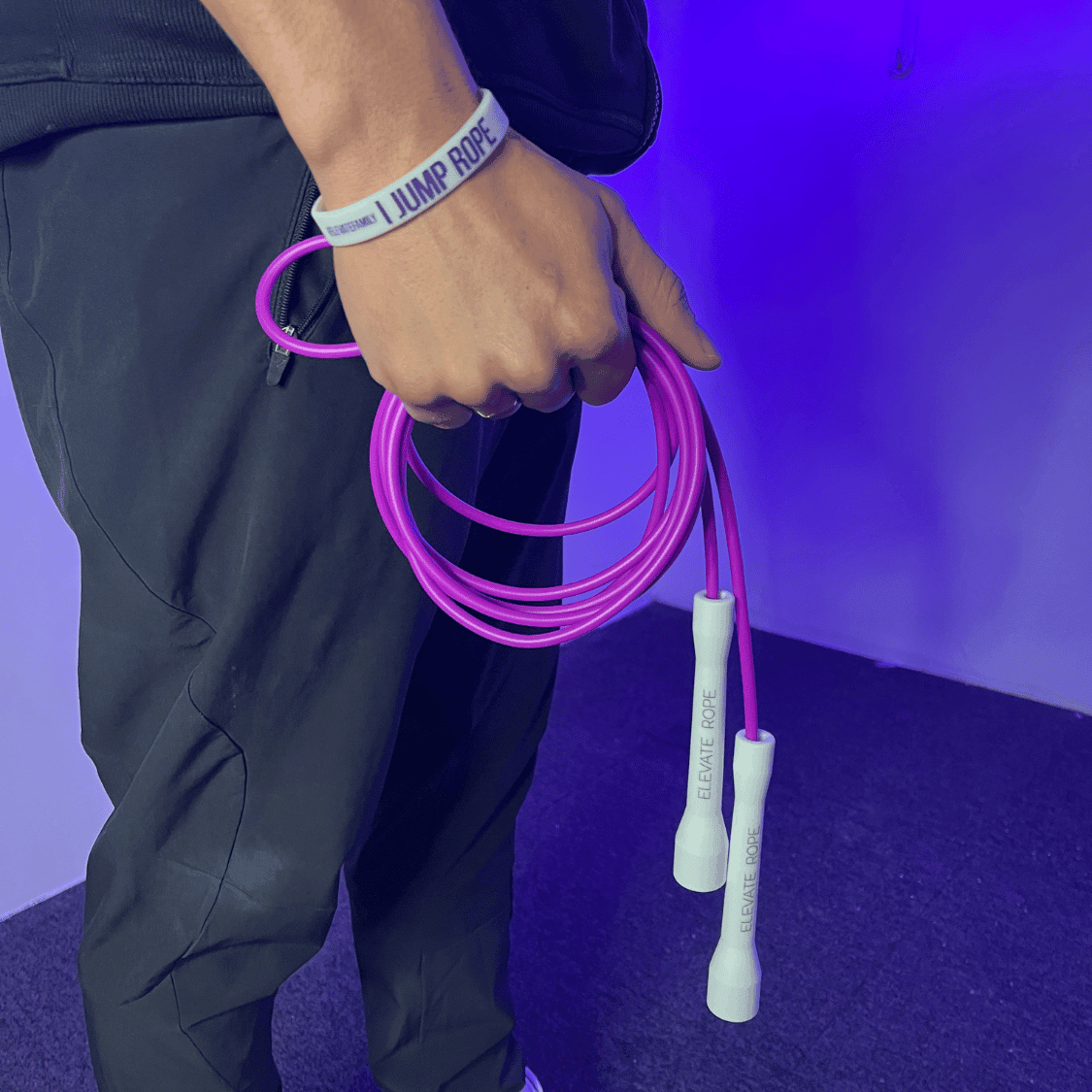 Purple jump rope with white handles, held by person wearing 'I Jump Rope' wristband, ideal for fitness workouts.